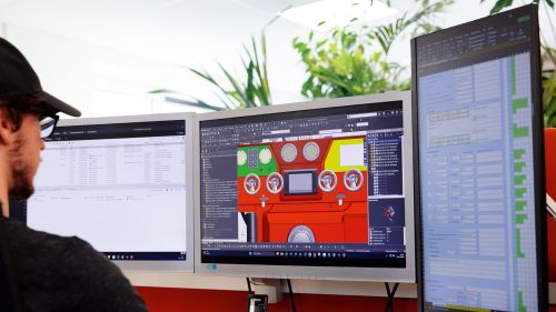 A person sitting in front of computer monitors, one of which displays a 3D design drawing