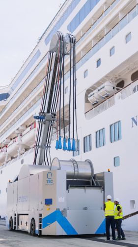 Several power cables are hanging down from the extended arm of the cable platform alongside a cruise ship.