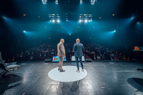 Bilgin Kilic und Björn Dunkel stehen auf einer runden, weißen Markierung auf der großen Bühne des Apollo Theaters vor gefüllten Zuschauerrängen. Sie halten ihren Vortrag „Mission Possible – The Clean Journey of Supply Chain Excellence“ bei den ifm SUCCESS DAYS 2026. Bilgin Kilic trägt einen hellen Anzug, Björn Dunkel einen dunklen Anzug und Sneaker. Im Hintergrund ist das Publikum im abgedunkelten Saal zu sehen.