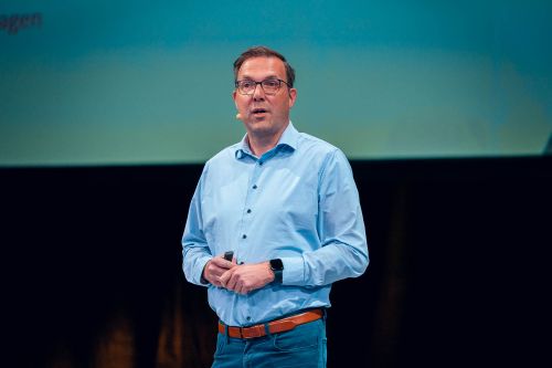 Steffen Brocksieper, Head of Production Logistics, hält einen Fachvortrag auf den Success Days. Er trägt ein hellblaues Hemd, Brille und ein Headset-Mikrofon, und hält einen Präsenter in den Händen.