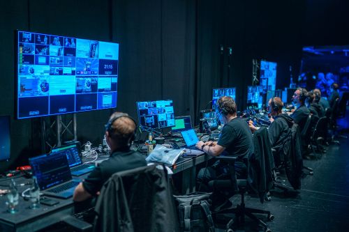 Ein atmosphärisches Foto aus dem Regie- und Backstage-Bereich der „Success Days“ im Apollo Theater. Im gedimmten Licht, dominiert von dunklen Wänden und Vorhängen, arbeitet ein Team von mehreren Veranstaltungstechnikern konzentriert an langen Tischen. Sie tragen schwarze Kleidung und professionelle Kommunikations-Headsets. Jeder Techniker bedient mehrere Laptops und Monitore, die die Live-Signale der Bühne, Grafiken und Regie-Multiview-Ansichten zeigen. An der linken Wand hängt ein sehr großer Hauptmonitor mit einem detaillierten Regie-Layout, das diverse Kameraeinstellungen und Textfelder für die Show-Ablaufsteuerung bereithält. Die Perspektive blickt über die Schultern des Technikers im Vordergrund auf die Reihe seiner Kollegen und die komplexe Licht- und Videotechnik, die einen reibungslosen Ablauf der Live-Show und des Streams sicherstellt.