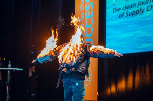Ein spektakulärer Stunt auf den ifm Success Days im Apollo Theater: Der Businessstuntman Holger Schumacher steht von hinten fotografiert mit ausgebreiteten Armen mitten auf der Bühne, während die Rückseite seiner Lederjacke und seine Ärmel lichterloh in Flammen stehen. Er trägt eine Jeans und ein schwarzes Shirt unter der brennenden Jacke. Im Hintergrund ist ein Teil der Veranstaltungsbühne zu sehen, darunter ein orangefarbenes Banner mit der Aufschrift „SUCCESS DAYS“ und eine große LED-Leinwand, auf der der Slogan „The clean journey of Supply Chain Excellence“ eingeblendet ist