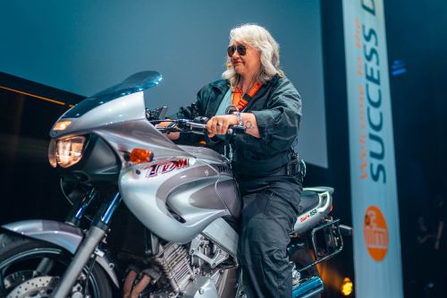 Ein dynamischer Auftritt auf der Bühne des Apollo Theaters während der ifm SUCCESS DAYS: Moderatorin Gerlinde Comisel sitzt im „Mission: Impossible“-Stil auf einem silbernen Yamaha TDM 850 Motorrad, das Scheinwerferlicht ist eingeschaltet. Sie trägt eine dunkle Fliegerjacke, eine passende Hose, eine Sonnenbrille und ein Headset-Mikrofon. Ihr Blick ist konzentriert nach vorne gerichtet. Im Hintergrund ist ein Teil der großen Bühne sowie rechts ein vertikales, weißes Banner mit dem ifm-Logo und dem Text „Welcome to the SUCCESS DAYS“ zu sehen.