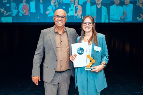 Bilgin Kilic von ifm und Linda Wismeth von der BHS Corrugated Maschinen- und Anlagenbau GmbH stehen lächelnd nebeneinander auf einer Bühne. Linda Wismeth trägt einen blaugrünen Hosenanzug und hält stolz die gläserne „ifm SCM Award 2026“-Trophäe sowie eine weiße Urkunde mit dem blauen ifm-Logo in den Händen. Bilgin Kilic, im grauen Sakko und braunen Hemd, steht links von ihr. Im Hintergrund ist eine große Leinwand zu sehen, auf der verschwommen weitere Personen mit Auszeichnungen abgebildet sind.