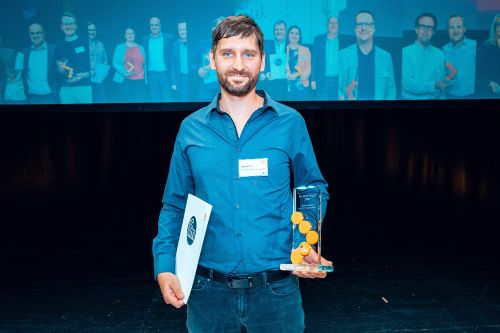 Bastian Graf von Gerresheimer steht lächelnd allein im Zentrum einer Bühne. Er trägt ein dunkelblaues Hemd und dunkle Jeans. In seiner linken Hand hält er die gläserne „ifm SCM Award 2026“-Trophäe mit den markanten orangefarbenen Kreisen, in seiner rechten Hand eine weiße Urkunde mit dem ifm-Logo. Im Hintergrund befindet sich eine große Leinwand, auf der im blauen Licht die anderen Preisträgerinnen und Preisträger der Verleihung eingeblendet sind.