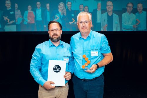 Martin Jansen und Armin Kurtzweg von der SMS group stehen lächelnd nebeneinander auf einer Bühne. Beide tragen hellblaue Hemden mit dem Logo der SMS group. Armin Kurtzweg (rechts) hält die gläserne „ifm SCM Award 2026“-Trophäe mit den markanten orangefarbenen Kreisen in der Hand. Martin Jansen (links) hält eine weiße Urkunde mit dem blauen ifm-Logo. Im Hintergrund ist eine große Leinwand zu sehen, auf der verschwommen die übrigen Gewinnerinnen und Gewinner der Verleihung abgebildet sind.