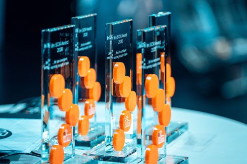 Fünf edle, gläserne Pokale der „ifm SCM Awards 2026“ stehen nebeneinander auf einem Tisch. Die transparenten Awards sind mit dem eingravierten Schriftzug „...for an excellent running supply chain“ versehen. Auf der Vorderseite jedes Pokals ragen die markanten, leuchtend orangefarbenen Kreise des ifm-Logos dreidimensional hervor. Der Hintergrund ist in stimmungsvollem, professionellem Blau gehalten.