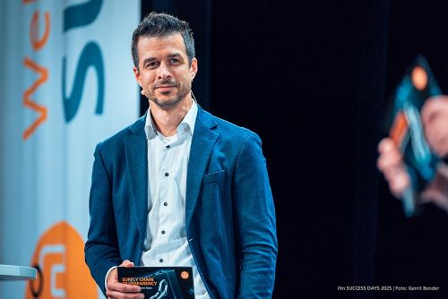 Foto von Alexander Römer in einem blauen Anzug und weißem Hemd, der bei den ifm SUCCESS DAYS 2025 lächelt und eine schwarze Broschüre mit dem Titel 